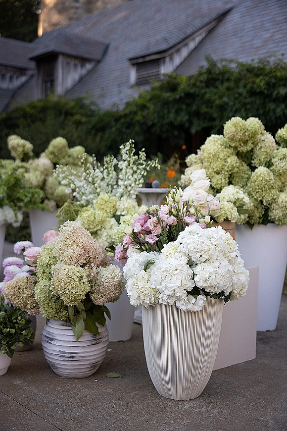 Wedding floral arrangements with hydrangea wedding florals in large white floor vases, featuring roses and greenery on an outdoor patio near a house exterior
