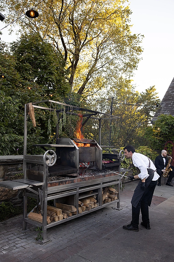 Wedding catering with a chef grilling over a live fire grill as flames rise from a metal station on a string-lit outdoor patio