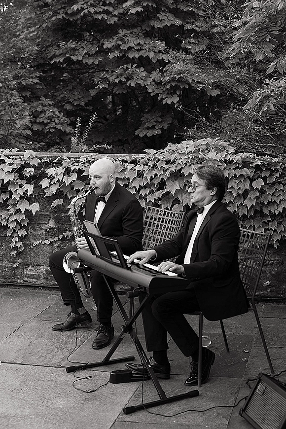 Wedding musicians in tuxedos play saxophone and keyboard with sheet music on an outdoor patio by an ivy-covered stone wall