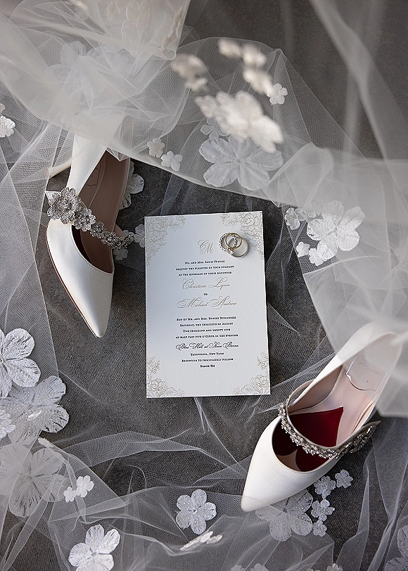 Wedding invitation flatlay with rings on the invitation, bridal heels and lace veil arranged on a gray fabric surface
