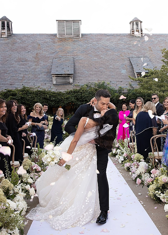 Wedding kiss portrait as the groom dips the bride on a white aisle runner, bouquet in hand amid rose petals and cheering guests in an ivy courtyard