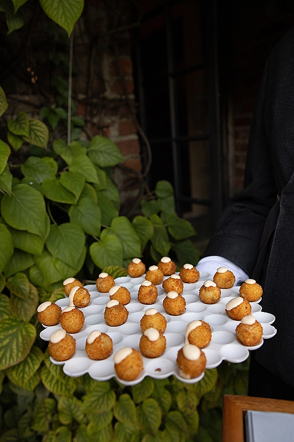 Wedding passed appetizers on a white serving tray with fried bites and dipping sauce held by a server, set against green foliage and brick wall