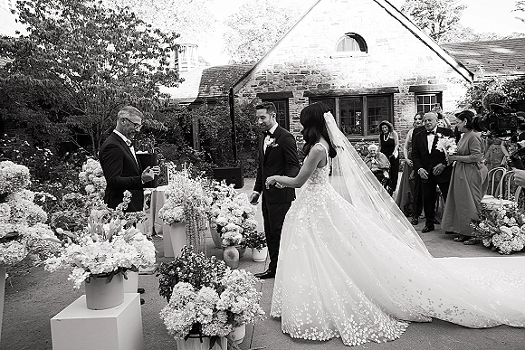 Outdoor ceremony moment as bride and groom at altar exchange vows, bride’s veil trailing, floral pedestals in a stone courtyard garden