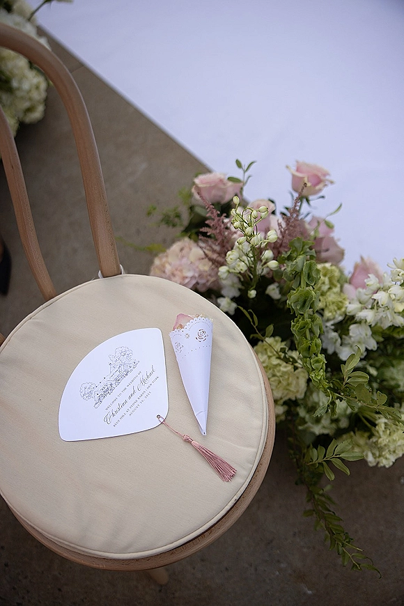 Ceremony programs with a confetti cone and pink tassel rest on a cushioned wood chair beside roses and hydrangea by the aisle runner