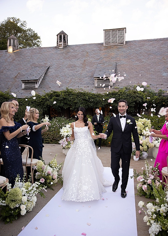 Wedding recessional as bride and groom walk the aisle holding hands, veil and bouquet visible, with rose petal toss on a white runner outside an ivy-covered building