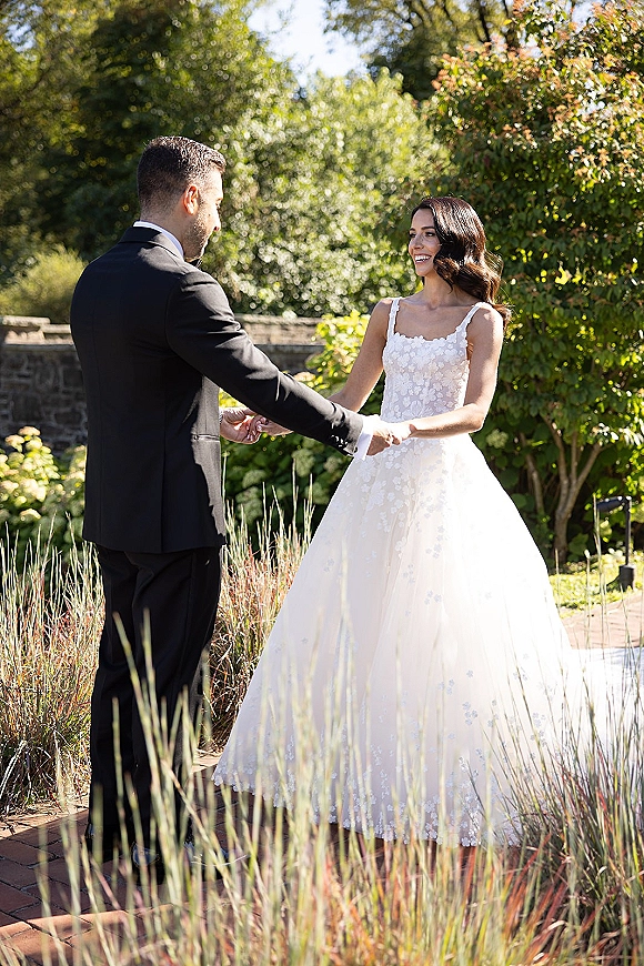First look moment as bride and groom holding hands on a brick path, bride in lace appliqué tulle gown, garden trees and stone wall behind