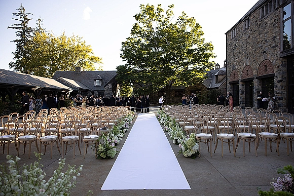 Ceremony aisle design with a white aisle runner flanked by low floral arrangements and wooden chairs in a stone courtyard with guests