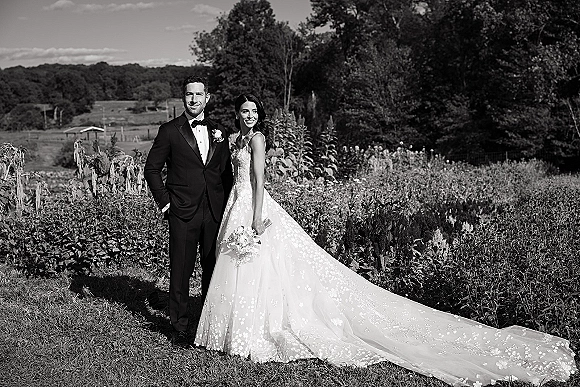 Couple portrait of bride holding bouquet beside groom in tuxedo, her lace wedding dress long train flowing in a wildflower field