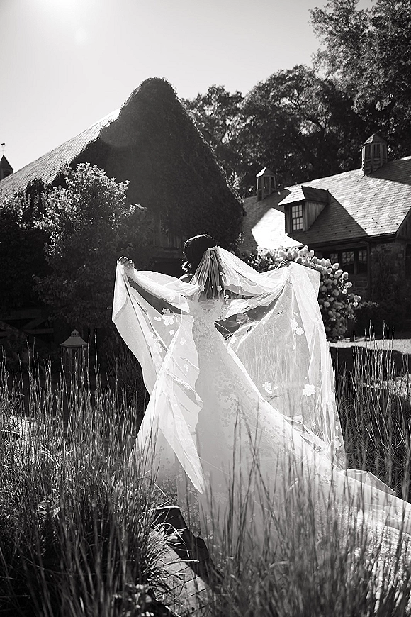 Bridal portrait of a bride back view in a strapless lace gown holding her floral applique veil, standing in tall grass by a sunlit stone house