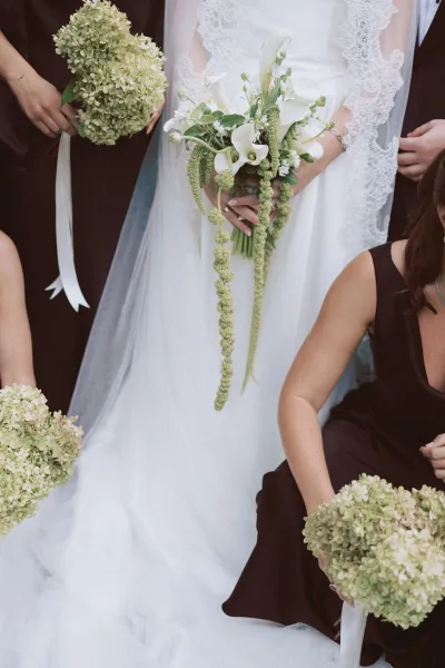 Bridesmaid bouquets of green hydrangea bridesmaid bouquets tied with white ribbon, held beside a lace-wrapped bride in a neutral indoor setting