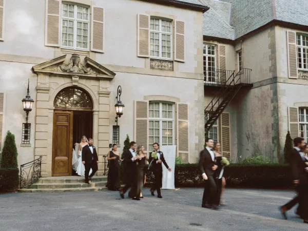 Wedding recessional as bride and groom exit ceremony with bridesmaids and tuxedo groomsmen, bouquet accent, from stone manor arched doorway lit by lanterns