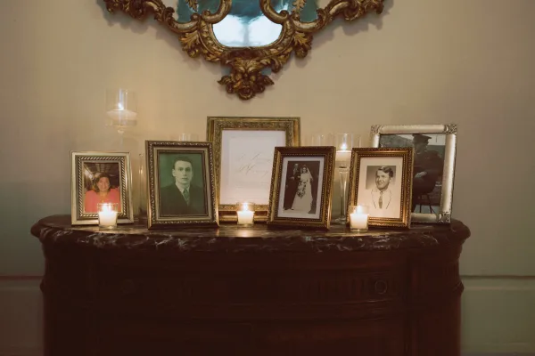 Memorial table with framed photos and candles in glass hurricane holders beside an ornate wall mirror, featuring a printed memorial sign
