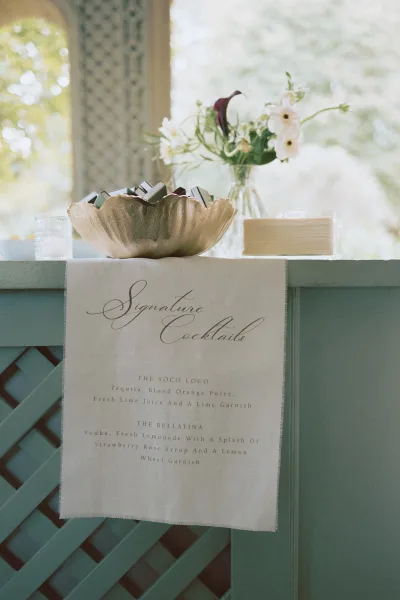 Signature cocktail sign with wedding signature drink menu in calligraphy on ribbon-edged paper, set by glassware and white florals outdoors