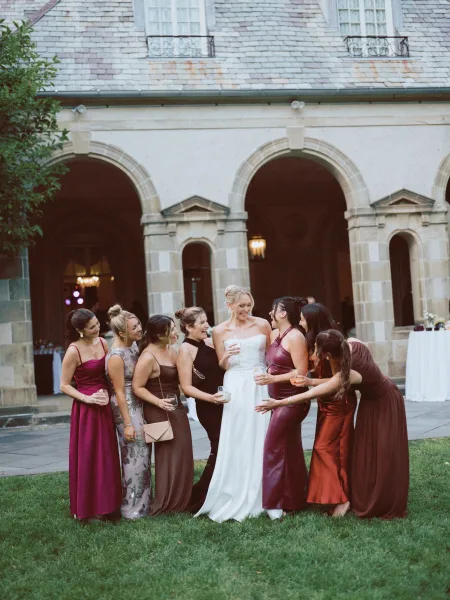 Bridesmaid group photo of bride with bridesmaids laughing, holding cocktail glasses in a courtyard by a stone archway with grass lawn trees