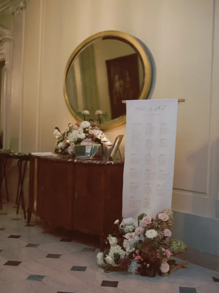 Seating chart display on a wooden console with floral arrangement and candles, framed photos and round mirror in an indoor entryway