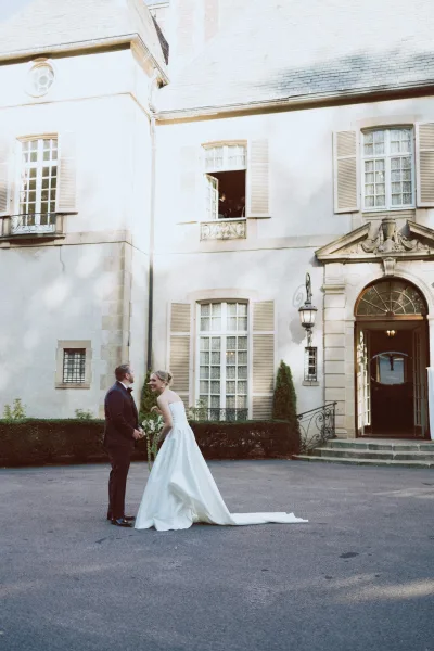 Couple portrait of bride and groom laughing, holding hands as she holds a green-and-white bouquet outside a mansion entrance on steps