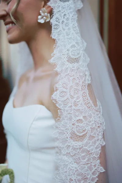 Bridal veil draped over the bride’s shoulder, lace wedding veil detail with pearl floral earrings beside a wooden door indoors