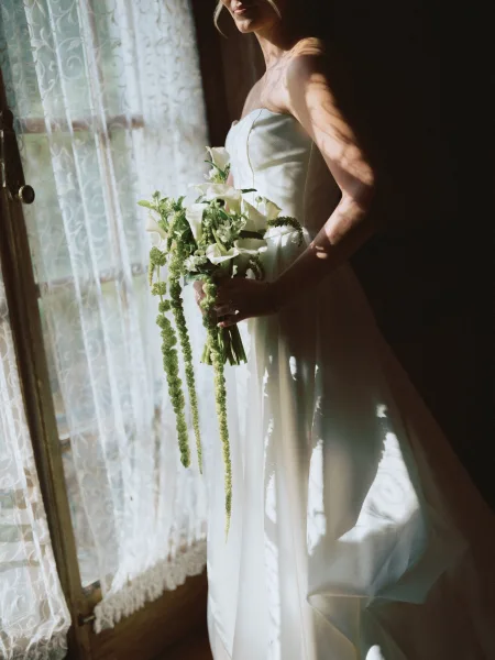 Bridal portrait of a bride holding bouquet of white calla lilies with trailing amaranthus, in strapless satin gown by a lace-curtained window