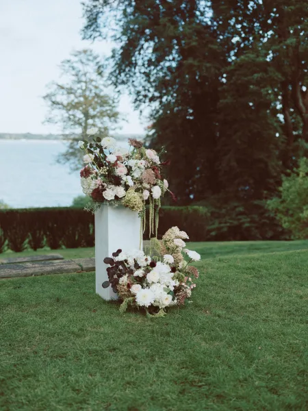 Ceremony floral arrangement of wedding ceremony flowers on a white pedestal with blush, burgundy, and white blooms, greenery, by a lake lawn