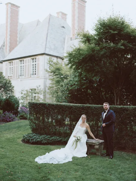 Couple portrait of bride in strapless dress with veil and bouquet seated on a stone bench, groom in tuxedo beside her in a manicured estate garden