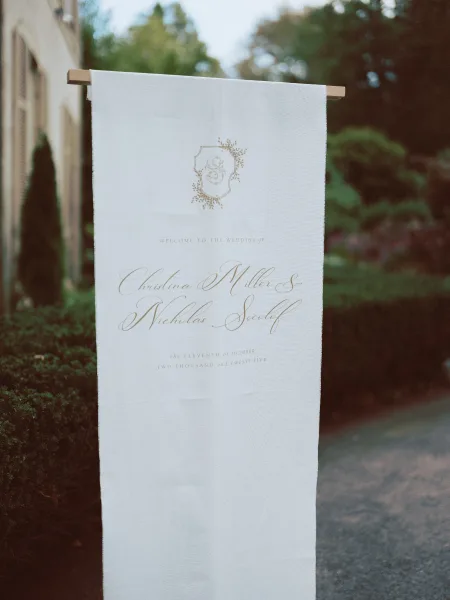 Wedding welcome sign with calligraphy lettering on a fabric wedding welcome sign, hanging from a wooden stand along a garden stone walkway