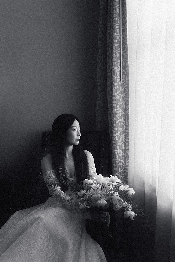 Bridal portrait in black and white of a bride seated by a window, holding a rose and greenery bouquet in an upholstered chair