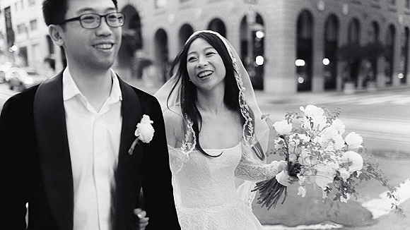 Wedding couple portrait of bride and groom walking, bride laughing with veil and bouquet beside an arched city street facade