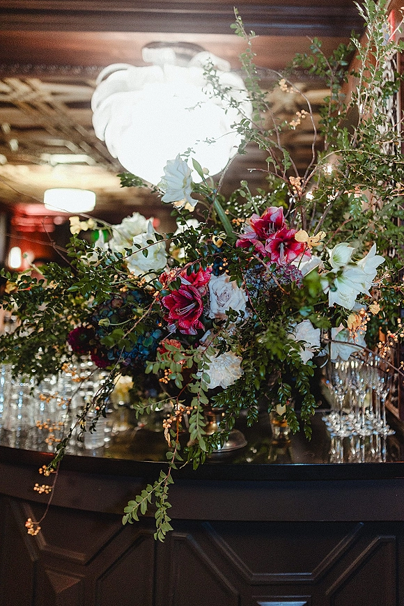 Wedding bar florals on the bar counter, a lush bar top floral arrangement with greenery, white, red and blue blooms beside champagne flutes