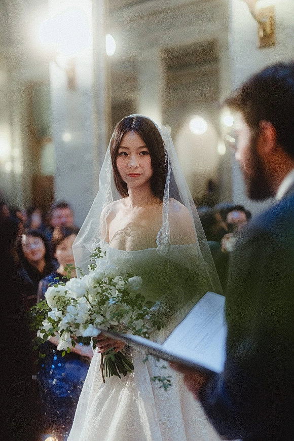 Ceremony moment as bride at altar holds a white rose bouquet in lace veil and dress, facing groom in dark suit under warm lights