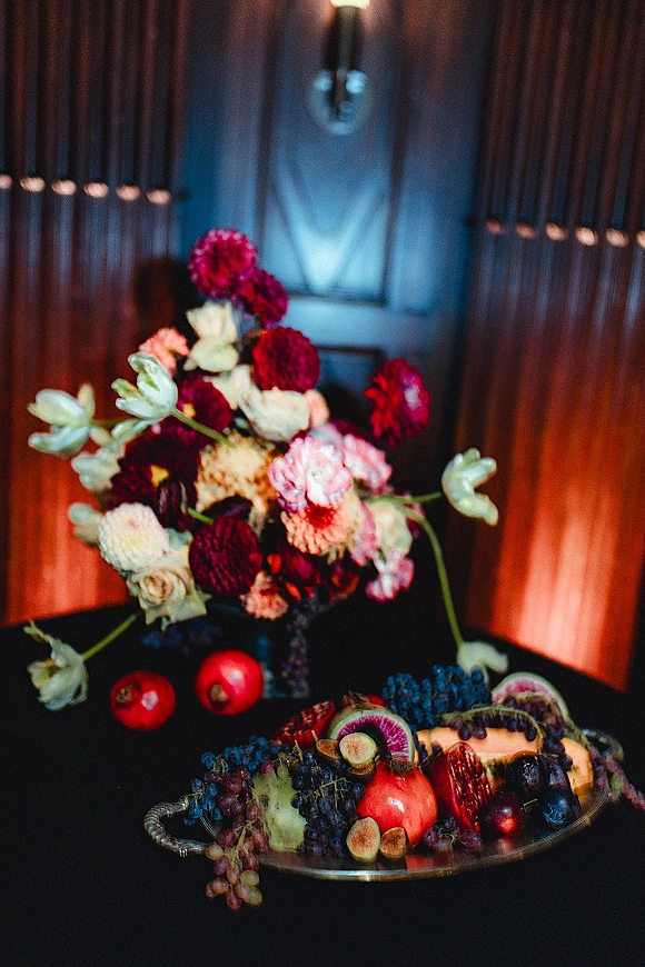 Wedding still life featuring a wedding fruit display with floral arrangement, grapes, pomegranates and citrus on a silver tray before a wood-paneled wall