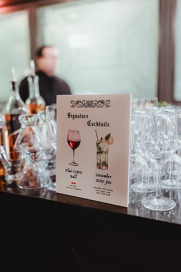 Signature cocktail sign listing wedding signature cocktails on a bar counter, with wine glasses and liquor bottles beside a blurred bartender