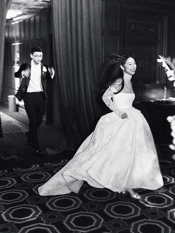 Wedding reception moment as bride runs laughing in lace-sleeved dress while groom in tuxedo chases her through dim indoor venue with draped curtains