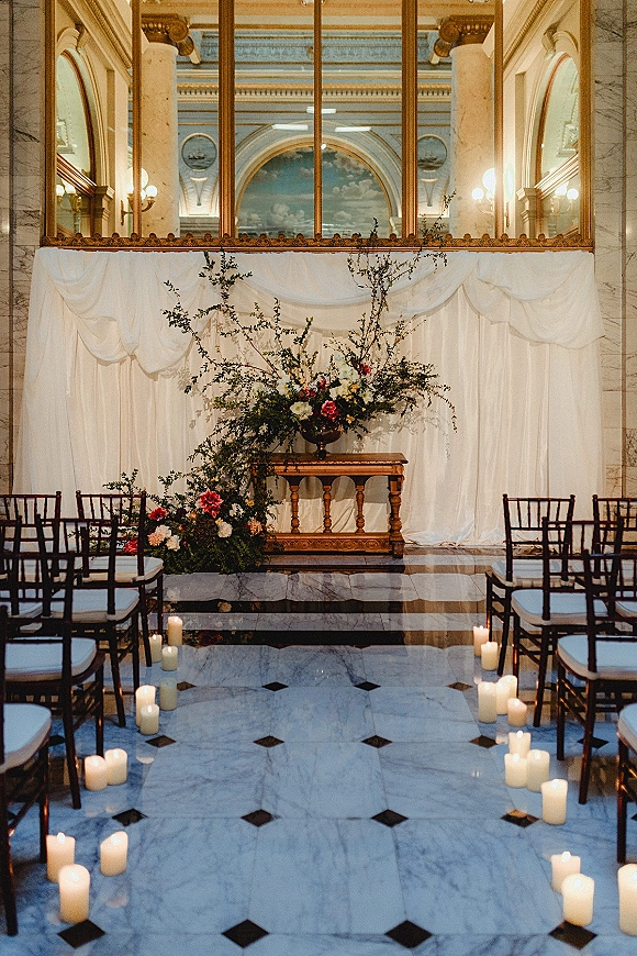Indoor ceremony setup with wedding ceremony backdrop draping, white fabric, greenery florals, pedestal altar table and candle-lined aisle in marble hall