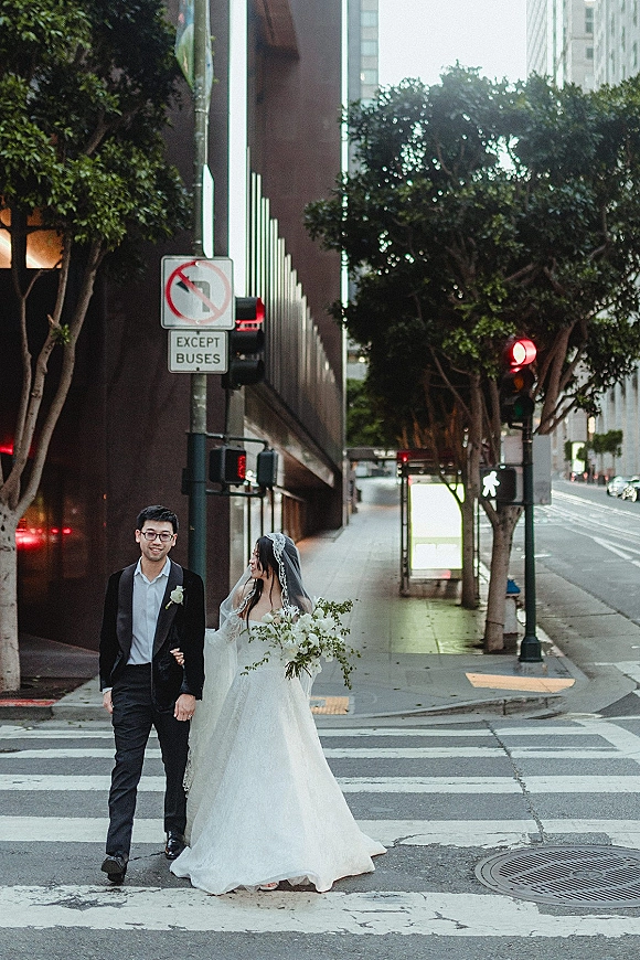 Couple portrait of bride and groom walking across a crosswalk, bride in veil holding bouquet beside tuxedoed groom downtown
