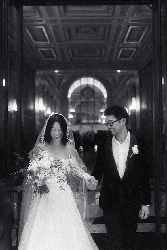 Couple portrait of bride in veil and lace dress holding a bouquet, walking hand in hand with groom in a coffered hallway