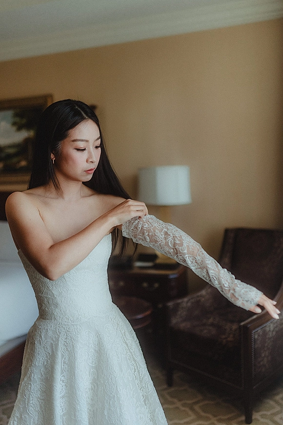 Bride getting ready in a hotel room, putting on lace sleeves over a strapless lace wedding dress, wearing simple earrings near the bed