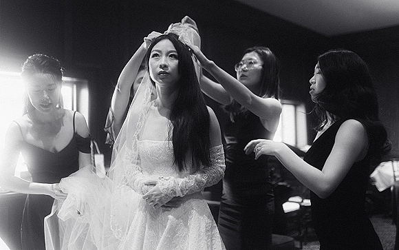 Bridal getting ready as bridesmaids help the bride putting on veil, her lace dress softly lit by window light in an indoor room