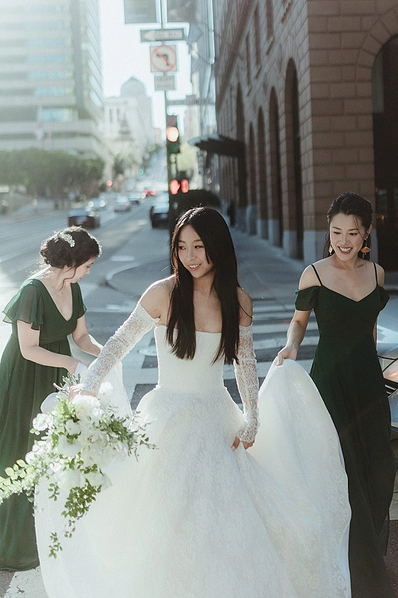Bride portrait walking a city street crosswalk holding a greenery bouquet, lace-sleeved gown as bridesmaids in dark green dresses lift her train