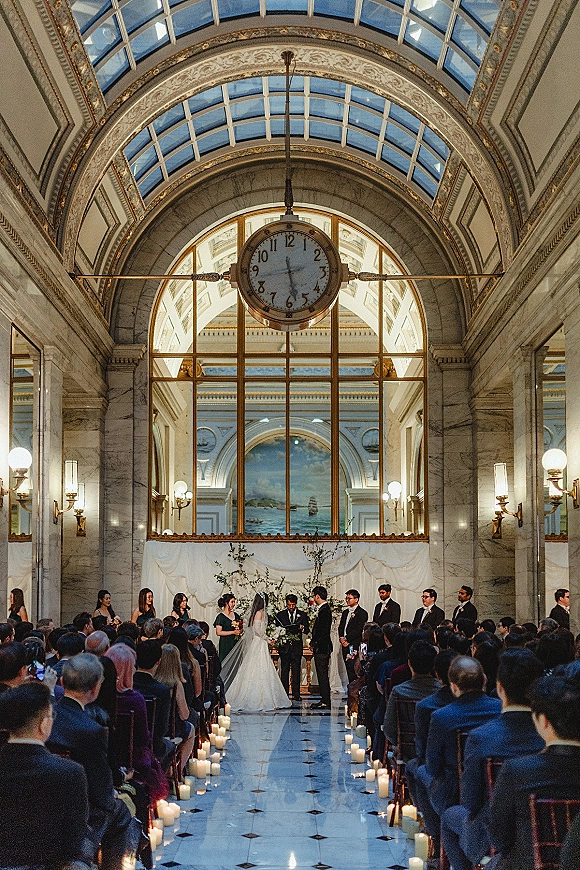 Wedding ceremony with couple at altar beneath a greenery arch, long veil and candlelit aisle in a grand marble hall with arched glass ceiling