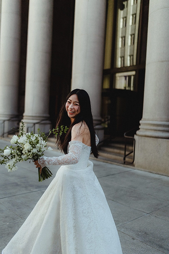 Bridal portrait of a bride holding bouquet, laughing as she looks back in an off-the-shoulder lace gown by stone columns