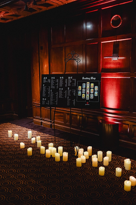Wedding seating chart on a black board easel with a floor of pillar candles and red uplighting against a wood-paneled wall