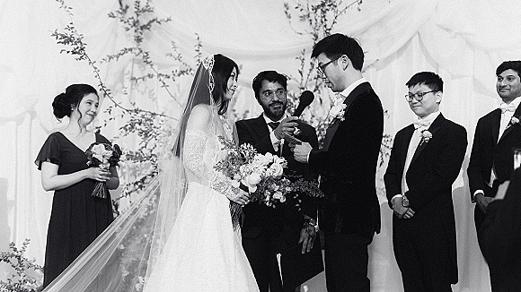 Wedding vows as groom reads from a card to bride holding bouquet with a cathedral veil, under draped fabric and greenery branches backdrop