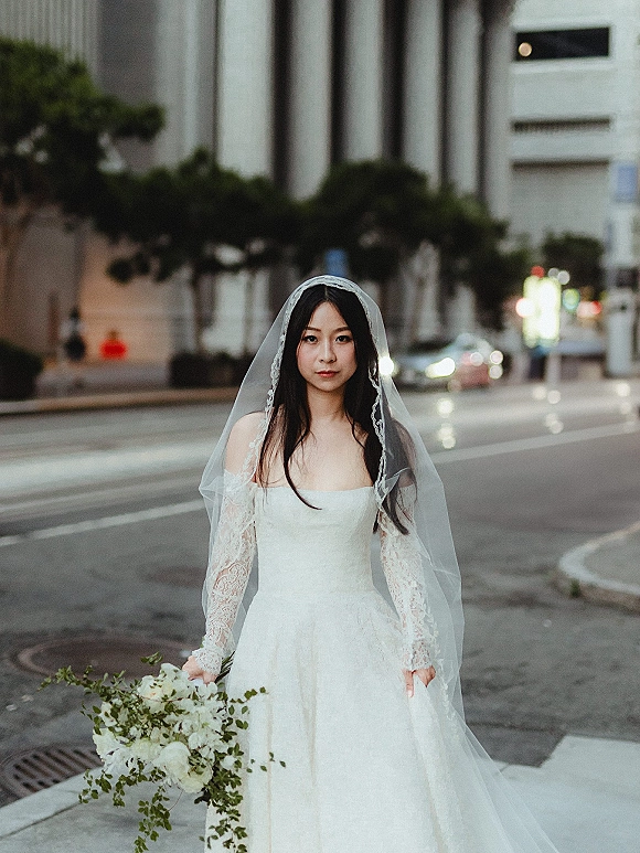 Bridal portrait of a bride in a lace-sleeve wedding dress and veil holding white flowers and greenery on a city street