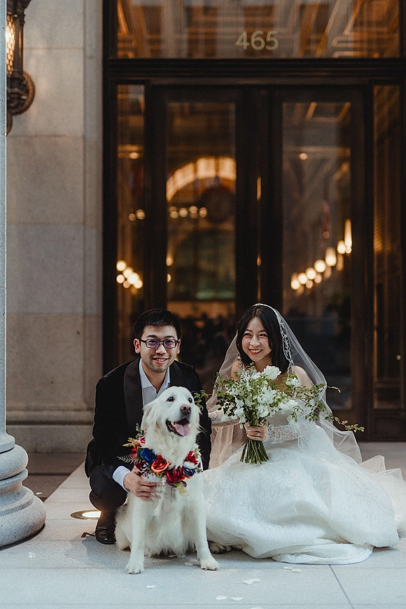Couple portrait of bride and groom with dog, bride in lace dress and veil holding white bouquet outside glass-door venue entrance