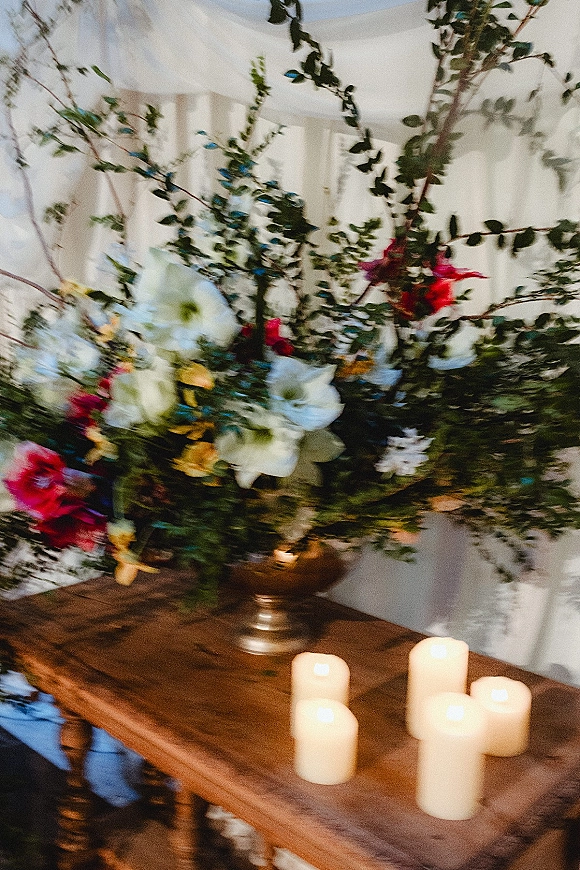 Wedding floral centerpiece in a silver urn vase with lush blooms, greenery branches, and pillar candles on a wooden table before a white paneled wall