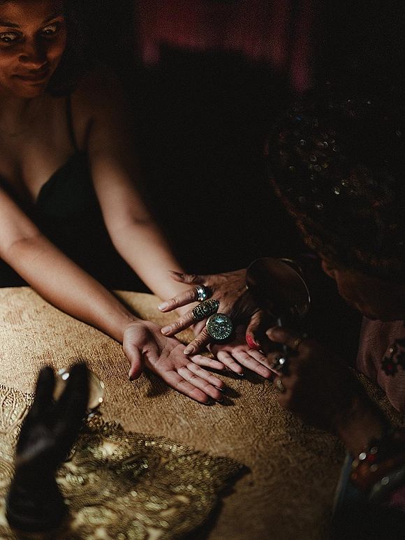 Palm reading at a wedding with hands resting on an embroidered pillow, statement rings and bracelets over a gold tablecloth in low light