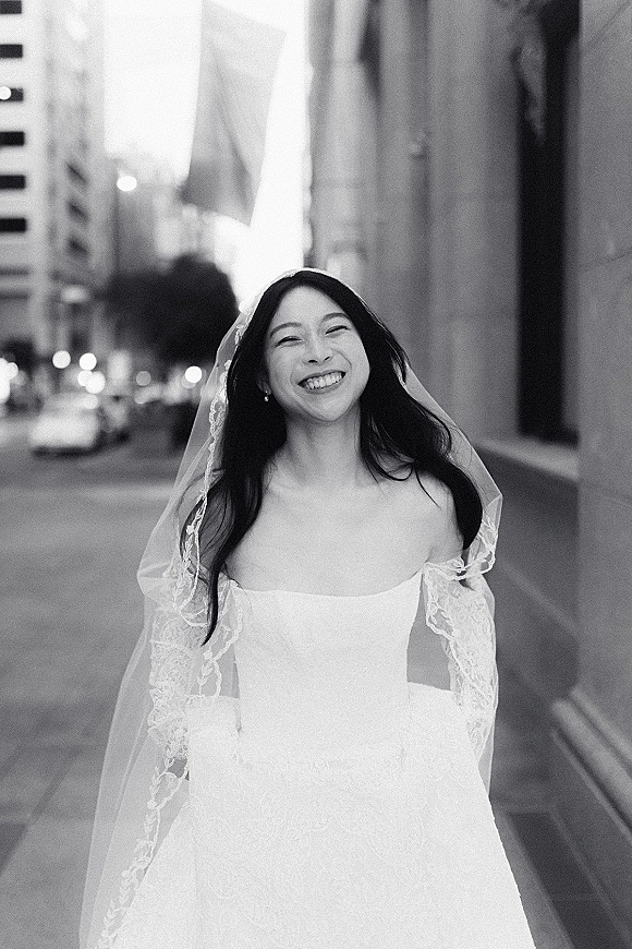 Bridal portrait of a smiling bride in a lace-sleeved wedding dress and veil walking along a city street with cars and trees behind