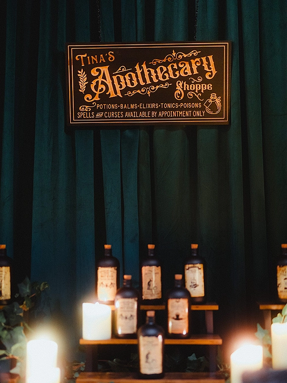 Wedding bar signage with signature cocktail sign hanging above glass bottles, pillar candles and greenery garland against a teal curtain backdrop