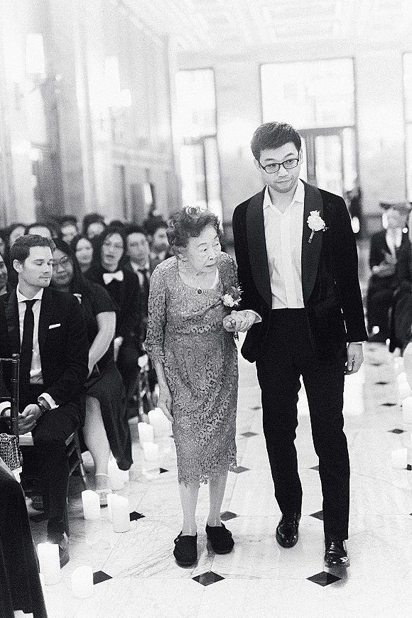 Wedding processional as groom walks down aisle with his grandmother, candle-lined marble floor and seated guests by large windows