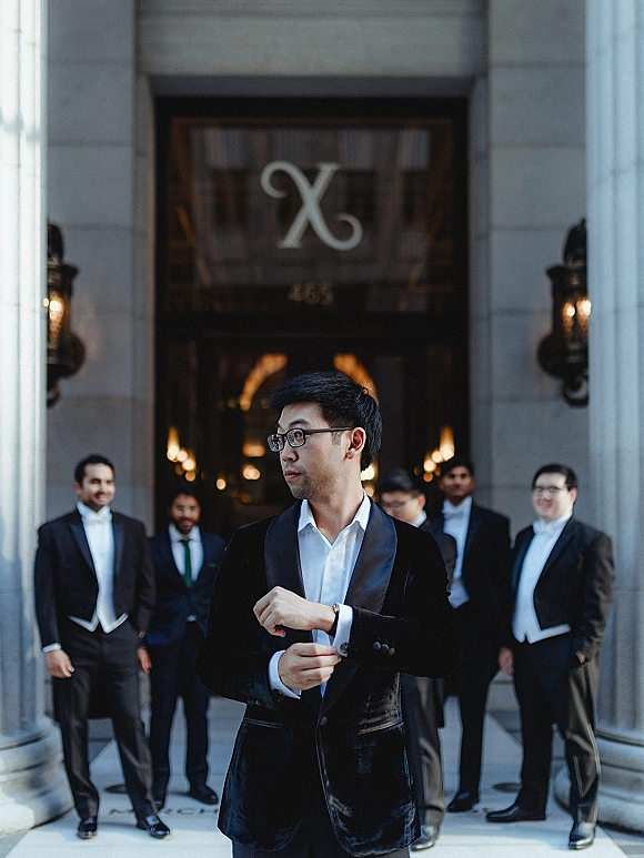 Groom portrait of a man adjusting cufflinks in a tuxedo with eyeglasses, standing at a building entrance with columns and sconces